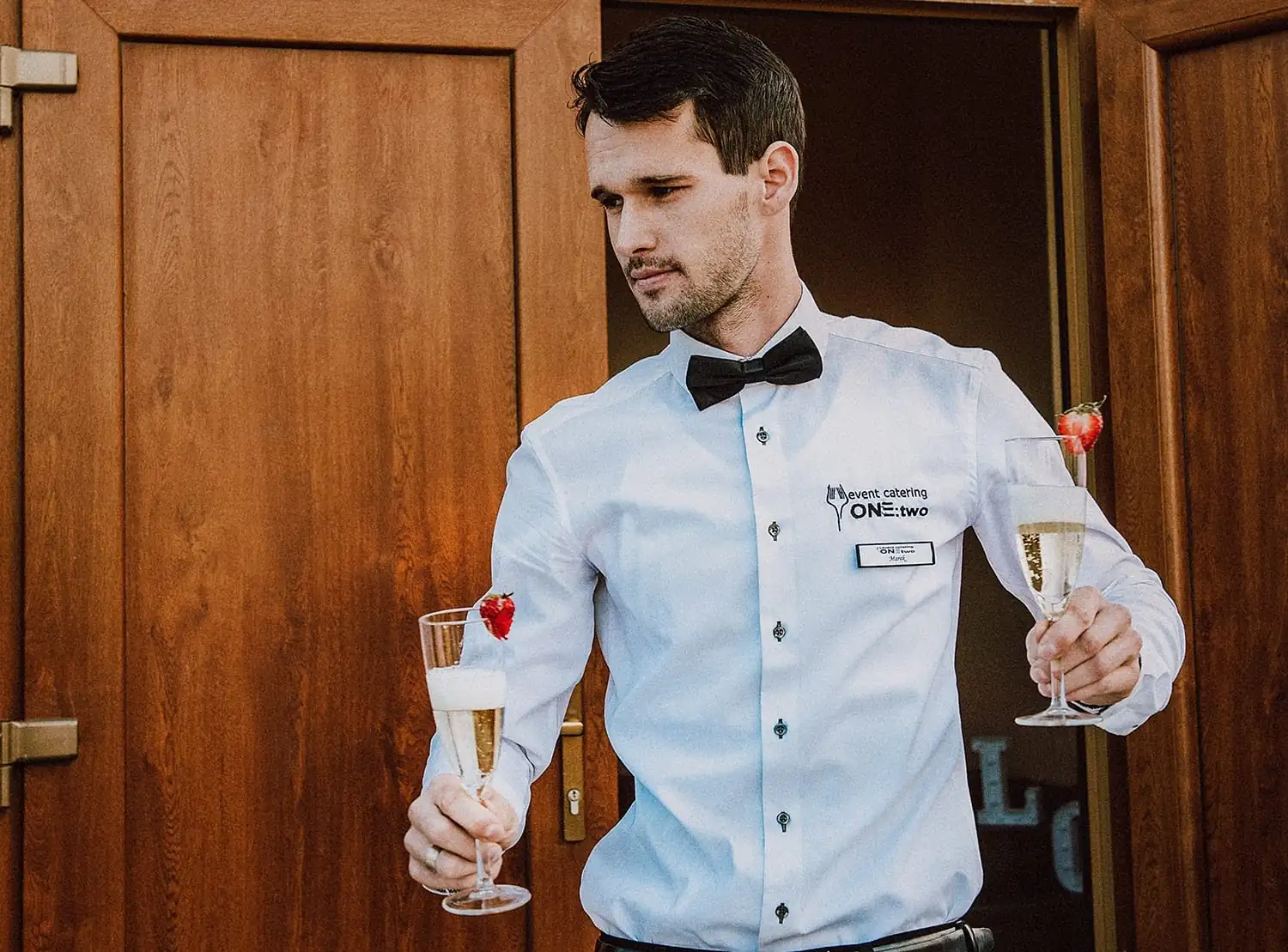 Ein Kellner im Hemd mit ONE:TWO-Logo bringt zwei Gläser Prosecco mit Erdbeere